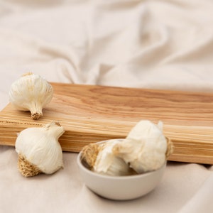 Puede incluir: Una tabla de cortar de madera con un patrón de rayas marrón claro y beige. Tres cabezas de ajo enteras y un cuenco blanco pequeño lleno de dientes de ajo están colocados sobre la tabla. El texto "THE WOODEN SURFACE" está impreso en la tabla.