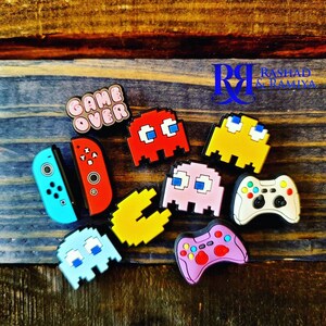 May include: A set of colorful Pac-Man and video game themed shoe charms, including a red Pac-Man ghost, a yellow Pac-Man ghost, a pink Pac-Man ghost, a blue Pac-Man ghost, a yellow Pac-Man, a red and black Nintendo Switch controller, a white Xbox controller, and a pink Xbox controller. The charms also include a pink and white "Game Over" charm.