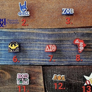 May include: A collection of colorful enamel pins with various Greek letters and symbols, including "ΦΒΣ", "ΩΨΦ", "ΖΦΒ", "ΔΣΘ", "ΚΑΨ", "AKA", and "ΑΦΑ". The pins are arranged on a wooden surface.