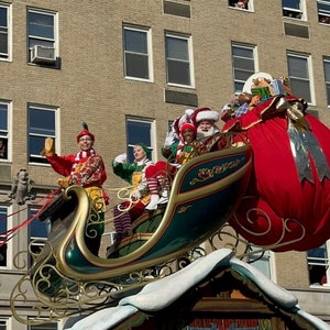May include: A festive red and green sleigh with Santa Claus and elves riding inside. The sleigh is pulled by reindeer and is decorated with lights and ornaments. The sleigh is flying through the air above a city street.