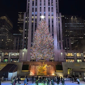 Può includere: Un grande albero di Natale decorato si erge di fronte all'edificio del Rockefeller Center a New York City. L'albero è illuminato con luci colorate e ornamenti. Le persone pattinano sul ghiaccio nella pista sotto l'albero.