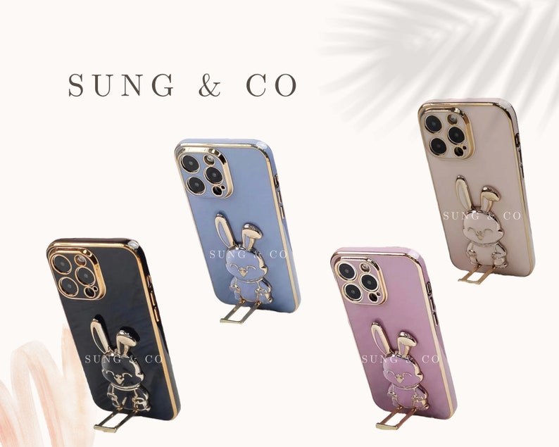 May include: Four phone cases with a gold trim and a 3D bunny design. The cases are in black, light blue, pink, and beige. Each case has the text "SUNG & CO" printed on the back.