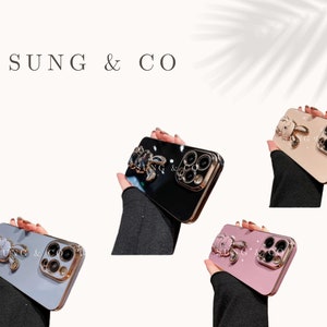 May include: Four iPhone cases with a decorative jeweled bunny charm attached to the back. The cases are black, white, pink, and beige.