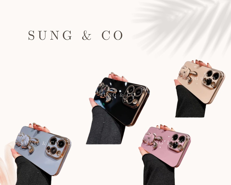 May include: Four iPhone cases with a decorative jeweled bunny charm attached to the back. The cases are black, white, pink, and beige.