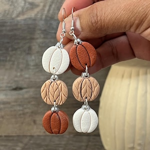 Puede incluir: Un par de pendientes colgantes con tres abalorios en forma de calabaza en blanco, beige y naranja. Los abalorios tienen una superficie texturizada y están conectados por anillos de salto plateados.