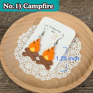May include: Campfire earrings with orange flames and yellow centers, resting on brown log bases. The earrings are 1.25 inches long and are displayed on a white card with the text "Designed by Siri". The text "No.1) Campfire" is in the top left corner.