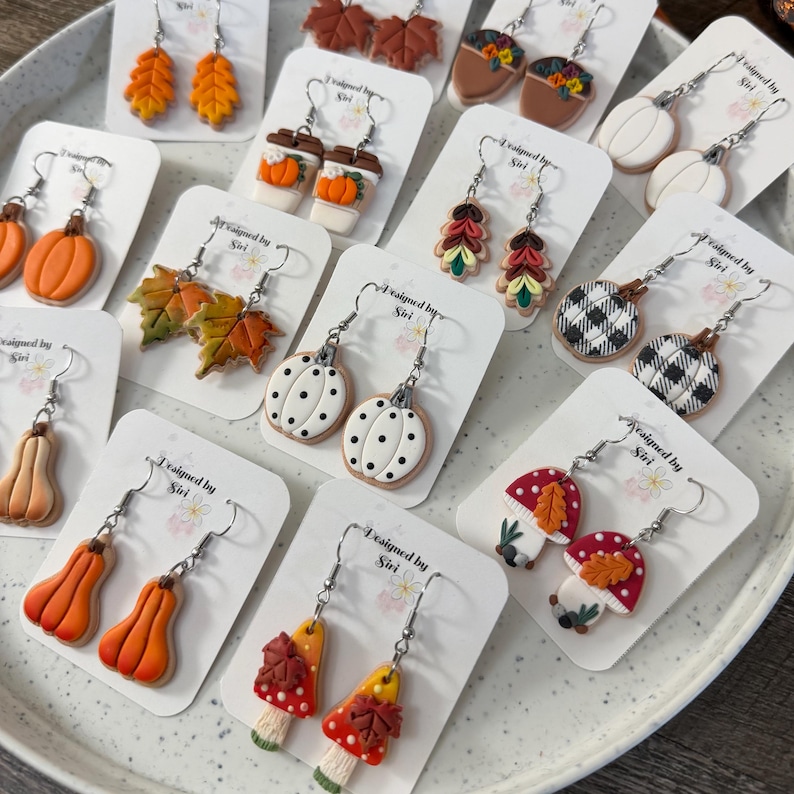 May include: A collection of polymer clay earrings on display cards, featuring autumn-themed designs including pumpkins, maple leaves, mushrooms, acorns, and coffee cups with orange, white, brown, and black patterns and textures.