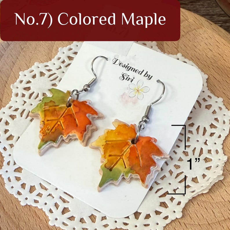 May include: A pair of dangle earrings shaped like maple leaves with a watercolor-style orange, yellow, and green finish. The earrings feature silver-tone metal hooks and measure 1 inch in length as indicated by the printed graphic.