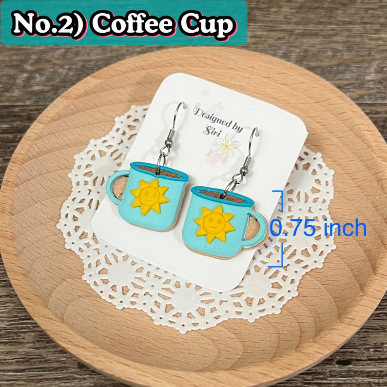 May include: A pair of blue coffee cup earrings with a yellow sun design. The earrings are 0.75 inches in length and are displayed on a white card with the text "Designed by Siri". The earrings are on a wooden plate with a white lace doily.