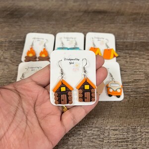 May include: A pair of cabin-shaped earrings with brown log walls, orange roofs, and yellow windows. The earrings are displayed on a white card with the text "Designed by Siri". Other earring designs include a campfire and a camper van.