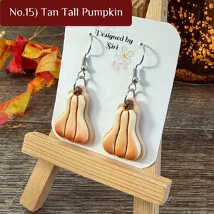 May include: A pair of dangle earrings featuring tan, tall pumpkin-shaped charms with brown stems. The earrings are attached to a white display card labeled No.15 Tan Tall Pumpkin and Designed by Siri with a flower icon.