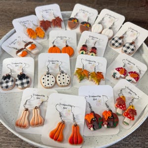 May include: A collection of polymer clay earrings displayed on white cards, featuring autumn-themed designs including pumpkins, maple leaves, mushrooms, acorns, and black and white buffalo plaid patterns, all attached to silver-toned metal earring hooks.