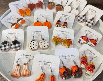 Pode incluir: Uma coleção de vários brincos pendentes com tema de outono exibidos em cartões brancos com a etiqueta Designed by Siri. Os brincos apresentam formas como abóboras, folhas de bordo, cogumelos, bolotas e padrões xadrez em laranja, branco e castanho.