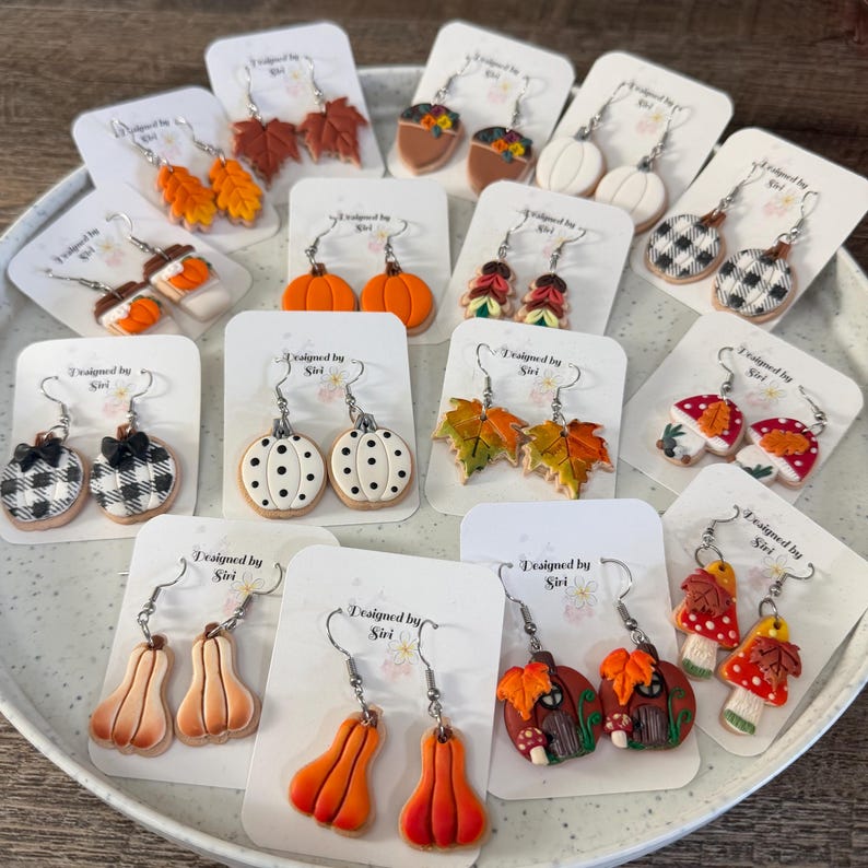 May include: A collection of various autumn-themed dangle earrings displayed on white cards labeled Designed by Siri. The earrings feature shapes including pumpkins, maple leaves, mushrooms, acorns, and buffalo plaid patterns in orange, white, and brown.