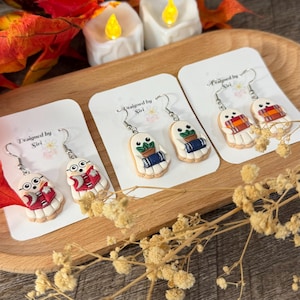 May include: Three pairs of white ghost-shaped earrings on white display cards labeled Designed by Siri. Each ghost features black eyes and holds different items, including a red book, a potted plant with a blue book, and a plaid scarf.