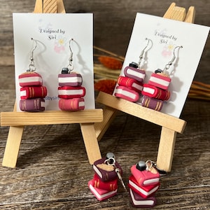 Puede incluir: Un par de pendientes con una pila de cuatro libros en miniatura con detalles dorados. Los libros son de color rojo, rosa y morado. Los pendientes están en un caballete de madera con un cartel que dice "Designed by Siri".