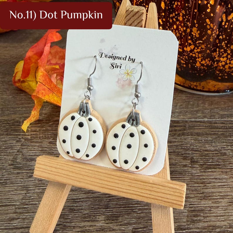 May include: A pair of dangle earrings featuring white pumpkin-shaped charms with black polka dots and grey stems. The earrings are displayed on a white card labeled No.11 Dot Pumpkin and Designed by Siri with a small flower icon.