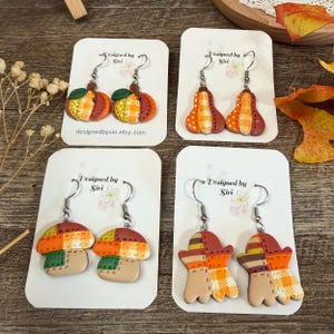 May include: Four pairs of dangle earrings on white display cards labeled Designed by Siri. The earrings feature clay shapes including pumpkins, gourds, mushrooms, and ghosts, all decorated with orange, yellow, and brown patchwork patterns.