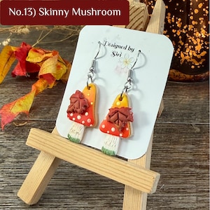 May include: A pair of dangle earrings featuring mushroom-shaped charms with orange and red caps, white spots, and a brown maple leaf accent. The earrings have silver-toned metal hooks and are displayed on a white card labeled No.13 Skinny Mushroom.