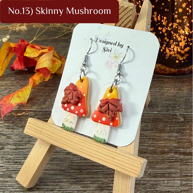 May include: A pair of dangle earrings featuring mushroom-shaped charms with orange and red caps, white spots, and a brown maple leaf accent. The earrings have silver-toned metal hooks and are displayed on a white card labeled No.13 Skinny Mushroom.