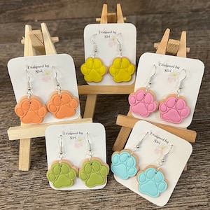 Puede incluir: Cinco pares de pendientes colgantes con dijes de huella de pata. Los dijes están hechos de arcilla polimérica y vienen en cinco colores: amarillo, naranja, rosa, verde y azul. Cada par de pendientes se muestra en un pequeño caballete de madera con una tarjeta que dice "Designed by Siri".