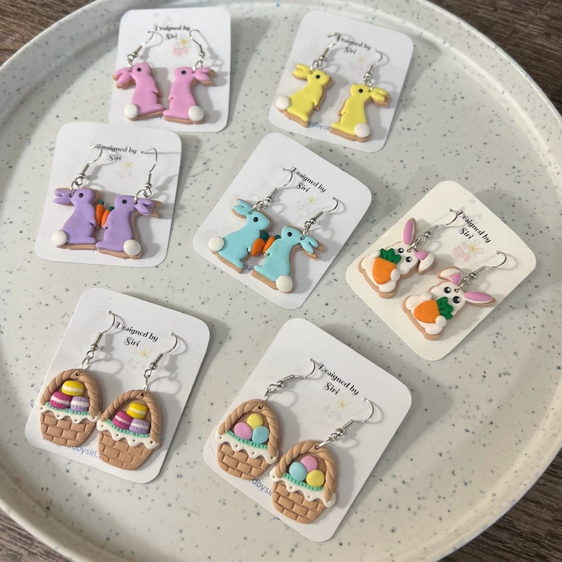 May include: Six pairs of handmade polymer clay earrings on white card stock with the text "Designed by Siri". The earrings feature Easter themes including bunnies, carrots, and baskets of eggs.