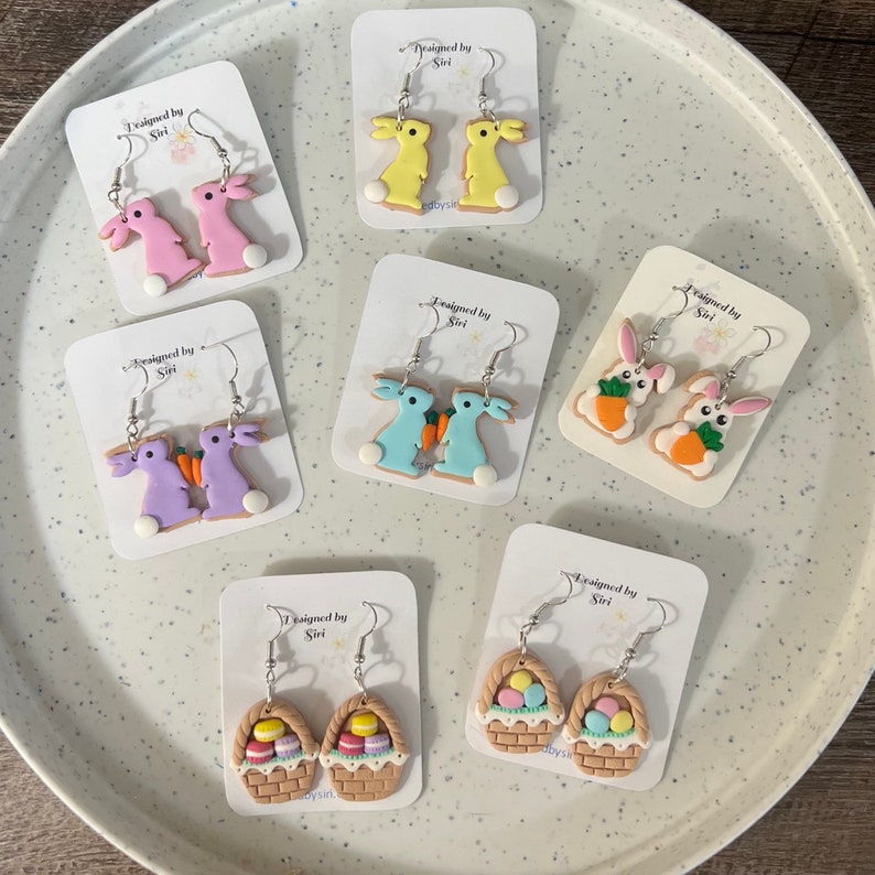 May include: Six pairs of handmade polymer clay earrings featuring Easter themes. The earrings are displayed on white cards with the text "Designed by Siri". The earrings include bunnies, carrots, baskets, and eggs.