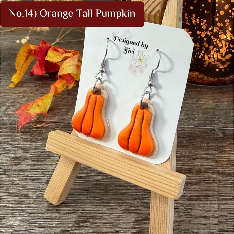 May include: A pair of dangle earrings featuring orange tall pumpkin charms with brown stems, attached to silver-toned metal hooks. The earrings are displayed on a white card labeled No.14) Orange Tall Pumpkin and Designed by Siri.