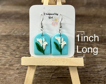 Pendientes colgantes de lirio de cala blanco sobre fondo azul de 1 pulgada de largo hechos de arcilla polimérica y alambres de acero inoxidable con forma de anzuelo. Idea de regalo para mamá.