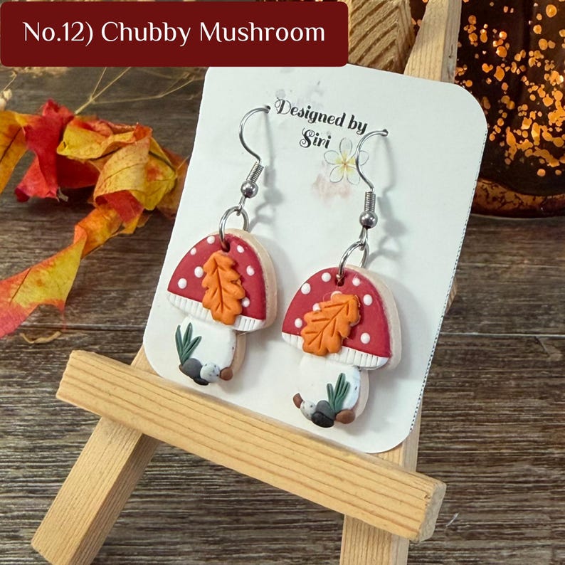 May include: A pair of dangle earrings featuring a red and white mushroom design with a small orange leaf accent on the cap. The earrings are displayed on a white card labeled No.12 Chubby Mushroom and Designed by Siri, featuring silver-toned hooks.