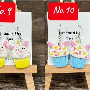 Może przedstawiać: Dwie pary ręcznie robionych kolczyk&oacute;w z polimerowej gliny. Pierwsza para przedstawia ż&oacute;łty cupcake z białym lukrem i r&oacute;żowymi uszami kr&oacute;liczka. Druga para przedstawia niebieski cupcake z białym lukrem i r&oacute;żowymi uszami kr&oacute;liczka. Obie pary mają posypkę na wierzchu. Kolczyki są oznaczone jako "No. 9" i "No. 10" i zostały zaprojektowane przez Siri.