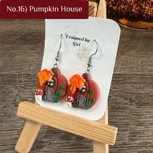 May include: A pair of dangle earrings featuring orange pumpkin-shaped charms with brown doors, windows, green vines, and orange leaves. The white display card reads No.16 Pumpkin House and Designed by Siri with a small flower illustration.