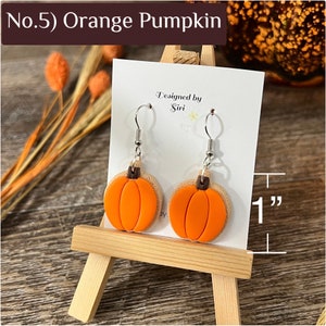 May include: A pair of orange pumpkin earrings with brown stems on silver-toned hooks, displayed on a white card labeled 'Designed by Siri'. A measurement graphic indicates the pumpkin charms are 1 inch in height.
