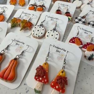 May include: A collection of polymer clay dangle earrings featuring autumn-themed designs including orange pumpkins, green and orange maple leaves, white pumpkins with black dots, red mushrooms with brown leaves, and black and white buffalo plaid pumpkins.