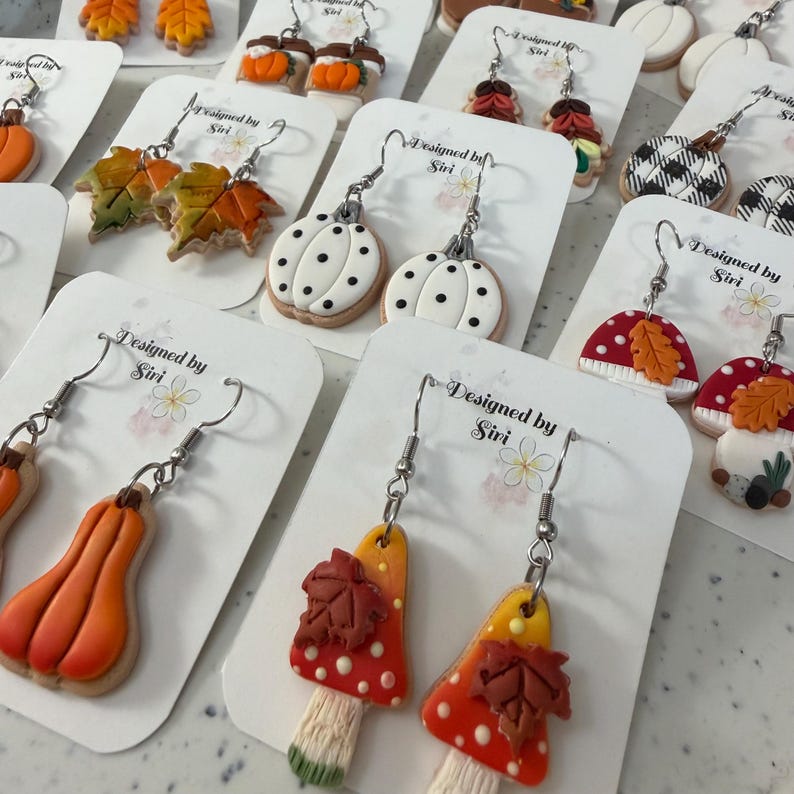 May include: A collection of polymer clay dangle earrings featuring autumn-themed designs including orange pumpkins, green and orange maple leaves, white pumpkins with black dots, red mushrooms with brown leaves, and black and white buffalo plaid pumpkins.