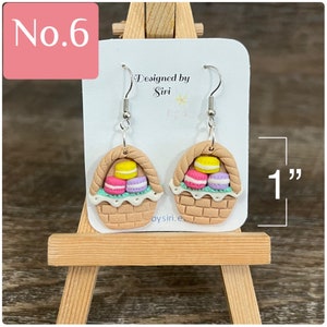 May include: A pair of brown and beige clay earrings shaped like baskets filled with pastel-colored macarons. The earrings are 1 inch long and have silver hooks. The earrings are designed by Siri.
