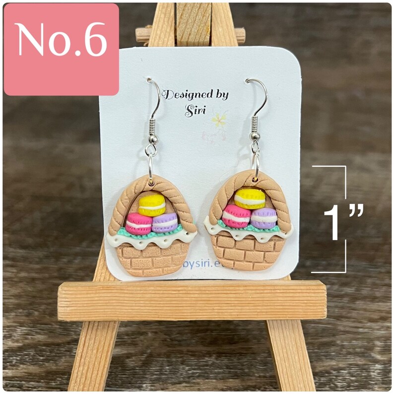 May include: A pair of brown and beige clay earrings shaped like baskets filled with pastel-colored macarons. The earrings are 1 inch long and have silver hooks. The earrings are designed by Siri.