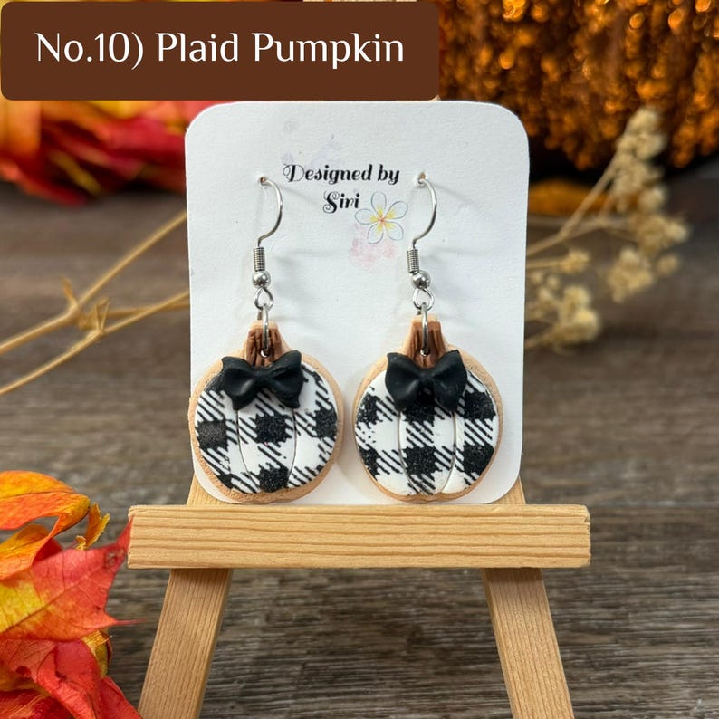 May include: A pair of dangle earrings featuring pumpkin-shaped charms with a black and white plaid pattern. Each pumpkin has a small black bow at the top and is attached to a silver-toned metal hook. The text No.10) Plaid Pumpkin is visible.