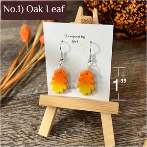 May include: A pair of dangle earrings featuring oak leaf charms with a yellow to orange gradient finish. The earrings hang from silver-toned hooks on a white display card labeled Designed by Siri. The leaf charms measure 1 inch in height.