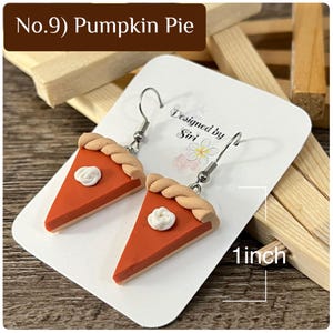 May include: A pair of pumpkin pie slice earrings with a tan crust, orange filling, and a white dollop of whipped cream. The earrings are displayed on a white card labeled No.9 Pumpkin Pie and Designed by Siri, showing a 1 inch measurement.