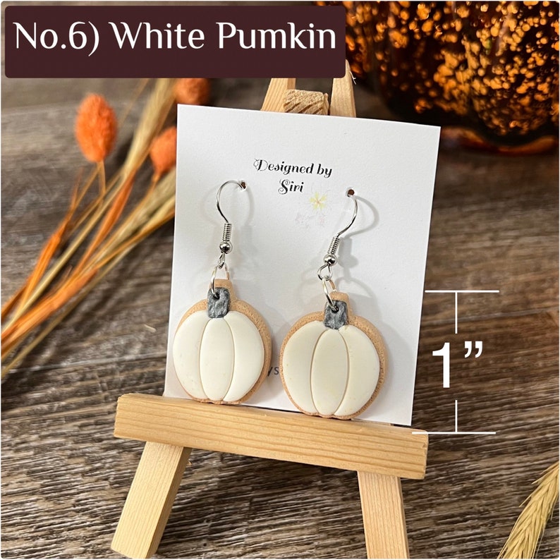 May include: A pair of white pumpkin-shaped dangle earrings with silver-toned hooks and gray stems. The earrings are displayed on a white card labeled 'Designed by Siri' and 'No.6) White Pumkin', with a graphic indicating a 1-inch height.