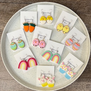 May include: Assortment of handmade polymer clay earrings with Easter themes.  The earrings feature designs such as chicks, rainbows, cupcakes, and eggs.  The earrings are displayed on white cards with the text "Designed by Siri".