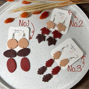 Könnte beinhalten: Drei Paar handgefertigte Ohrringe werden auf einem weißen Teller präsentiert. Die Ohrringe weisen verschiedene herbstliche Designs auf, darunter Blattformen und abstrakte Formen in Braun-, Beige- und Rottönen. Jedes Paar ist an einer weißen Karte befestigt.