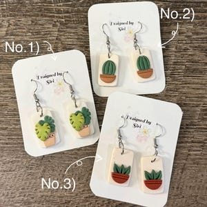 Peut inclure: Boucles d'oreilles faites à la main avec des motifs de plantes miniatures. Chaque boucle d'oreille présente une base rectangulaire blanche avec une petite plante détaillée dans un pot de couleur terre cuite. Le texte "Designed by Siri" est imprimé sur la carte.