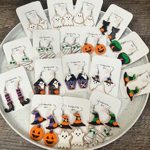May include: A collection of Halloween-themed earrings, including ghosts, pumpkins, witch hats, mummies, and haunted houses. The earrings are in various colors, including orange, black, white, and purple. Each pair is displayed on a white card.