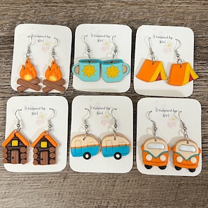 May include: A collection of handmade earrings featuring camping and outdoor themes. Designs include campfires, mugs with suns, orange tents, log cabins, blue and white campers, and orange vans. Each pair is attached to a white card with the text "Designed by Siri".