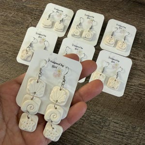 Könnte beinhalten: Handgefertigte, cremefarbene Ohrringe mit Muschel-, Spiral- und Seestern-Designs. Jeder Ohrring ist an einem silbernen Haken befestigt und auf einer weißen Karte präsentiert. Die Karten sind mit "Designed by Siri" beschriftet.