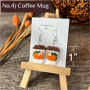 May include: A pair of dangle earrings shaped like coffee cups with brown lids and white bodies. Each cup features an orange pumpkin and a small white flower. The earrings measure 1 inch in height as indicated by the visible measurement graphic.