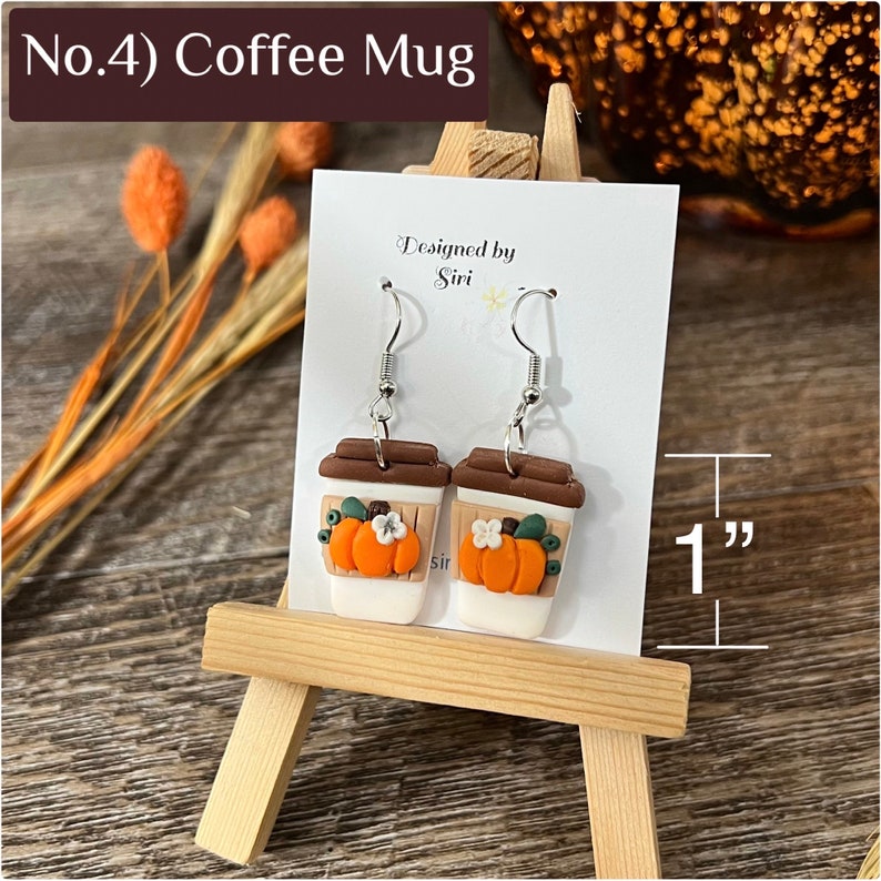 May include: A pair of dangle earrings shaped like coffee cups with brown lids and white bodies. Each cup features an orange pumpkin and a small white flower. The earrings measure 1 inch in height as indicated by the visible measurement graphic.