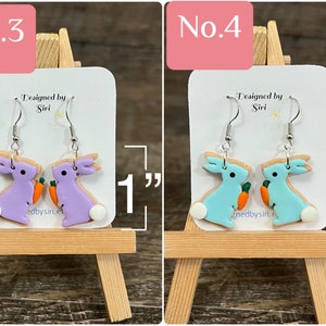 May include: Two pairs of dangle earrings featuring a bunny holding a carrot. The bunny on the left is purple and the bunny on the right is light blue. The earrings are 1 inch long. "Designed by Siri" is written on the card behind the earrings.